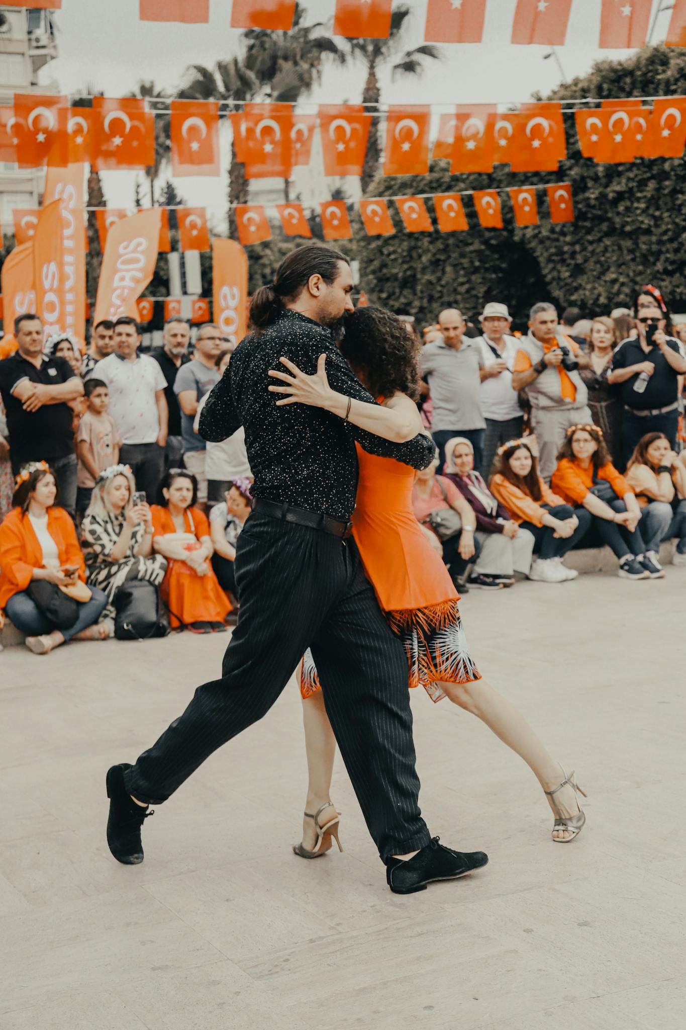 A couple performs a dance amidst a lively festival with an audience and Turkish flags.