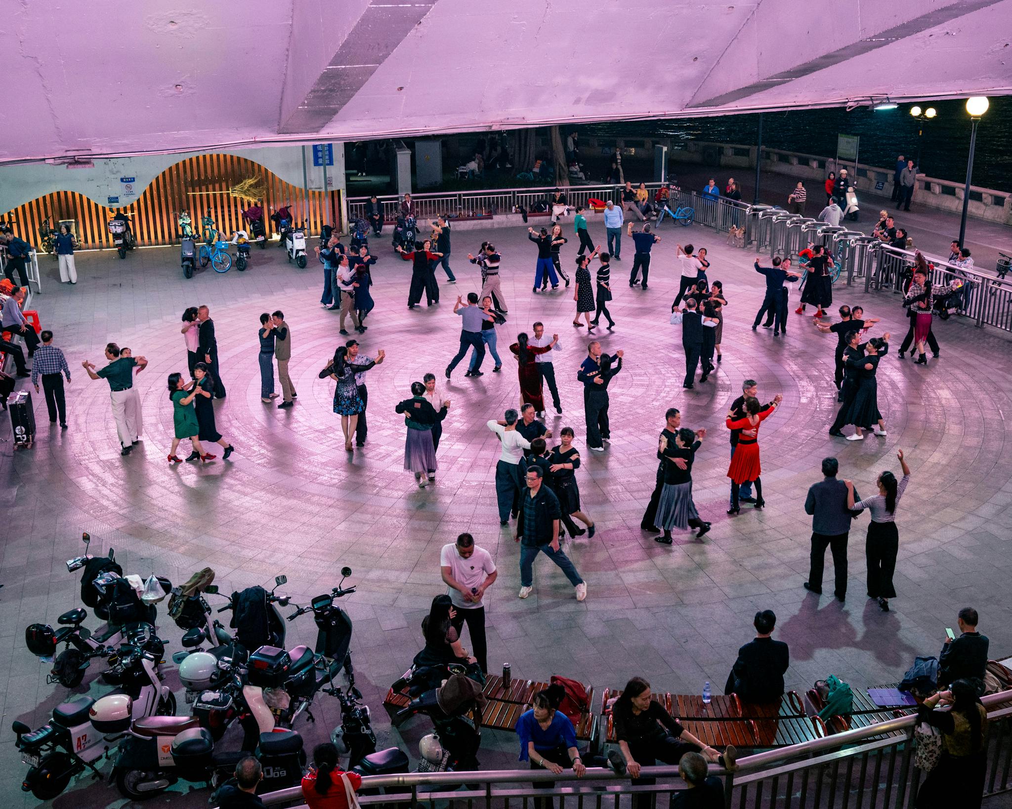 A large group of people dancing in an outdoor urban plaza under purple lights during the evening.